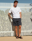 Man wearing black slide sandals, white t-shirt and dark shorts standing in front of a white picket fence.