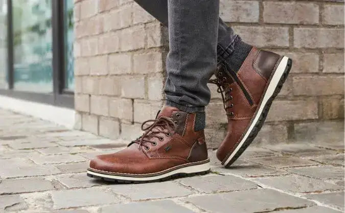Brown leather ankle boots with white soles and black treads, worn with dark jeans on a cobblestone sidewalk near a brick wall.