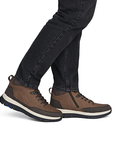 A person wearing brown mid-top lace-up shoes with dark jeans, shown from the calves down.