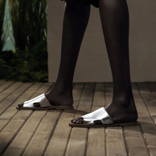 Model wearing Women's Bueno Hendrix Slide Sandal in silver leather, styled with dark tights on a wooden floor, showcasing its sleek flat design.