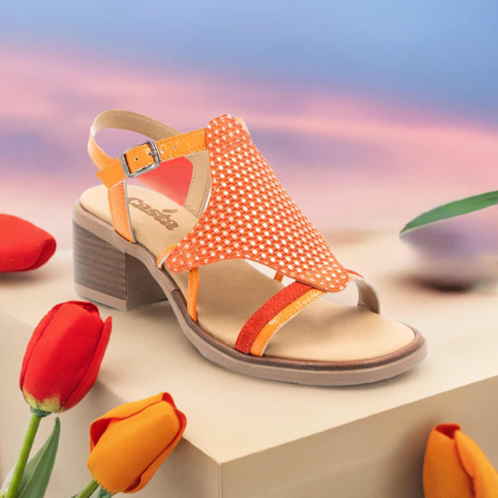 Women's Casta Cade Heeled Leather Sandal displayed among tulips, featuring vibrant orange leather, woven design, and comfort heel, handmade in Turkey.