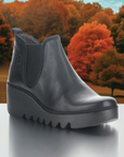 Women's FLY London Byne Wedge Boot in black leather displayed against a fall foliage background, ideal for autumn fashion and comfort.