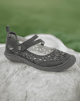 JBU Bellerose Encore Mary-Jane Flat in dark gray displayed outdoors on a rock, showcasing its floral perforations and rugged design.