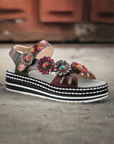 Women's L'Artiste Laga backstrap sandal displayed on rustic background, highlighting colorful floral leather and platform sole.