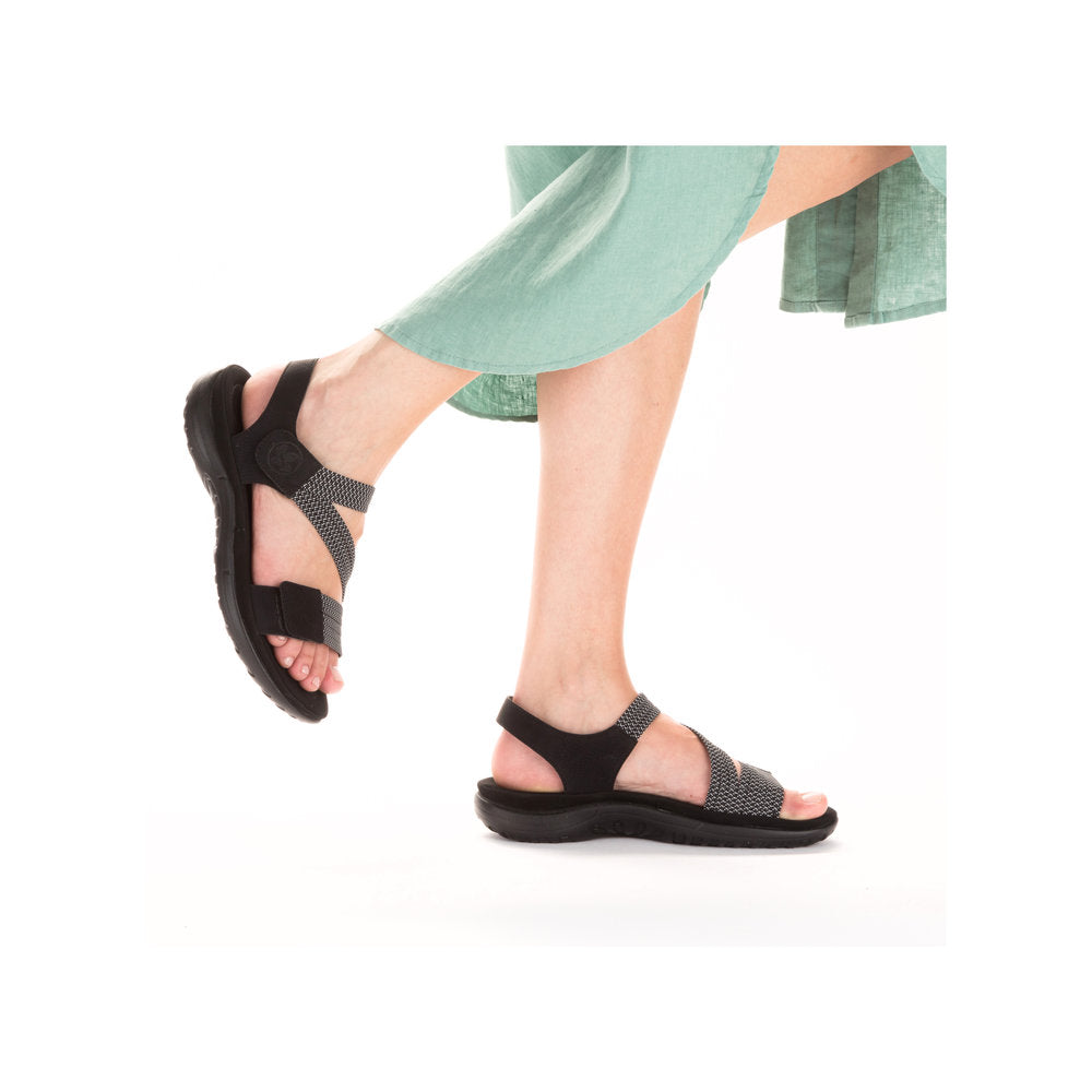 Model wearing black Women's Rieker 64870 Backstrap Sandals with a green dress, showcasing flexible fit and casual elegance.