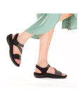 Model wearing black Women's Rieker 64870 Backstrap Sandals with a green dress, showcasing flexible fit and casual elegance.