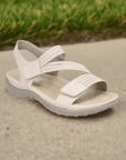 Beige Women's Rieker 64870 Backstrap Sandal displayed outdoors on pavement, highlighting its casual style and comfort fit.