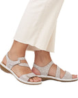 Model wearing white crackle Rieker 659C7 sandals with Velcro straps and cushioned sole, styled with cream pants.