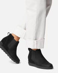 Model wearing black Women's Sorel Evie Pull On Wedge Boots with white pants, showcasing the ankle height and sleek waterproof design.
