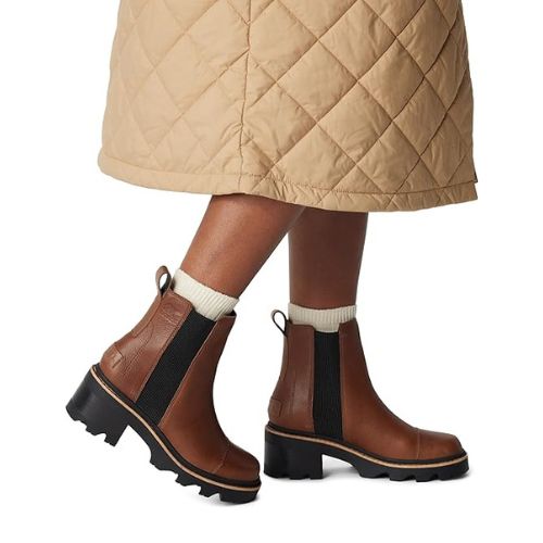 Model wearing Women's Sorel Joan Now Chelsea Waterproof Boots styled with a quilted skirt, highlighting ankle-height design and comfort for urban wear.