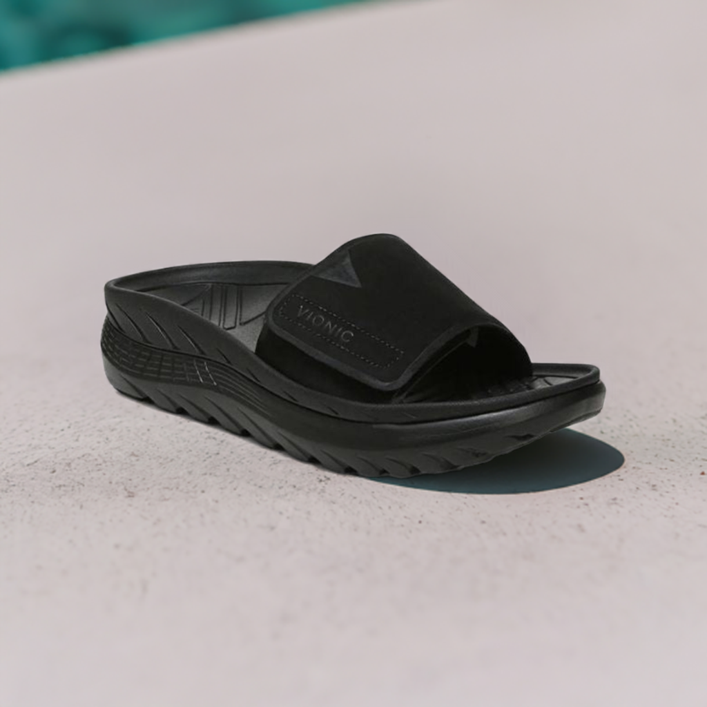 Vionic Rejuvenate Recovery Slide Sandal displayed outdoors, highlighting its sleek black design and supportive structure for post-activity wear.