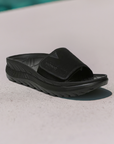 Vionic Rejuvenate Recovery Slide Sandal displayed outdoors, highlighting its sleek black design and supportive structure for post-activity wear.
