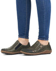 Woman wearing blue jeans and green slip on shoes.