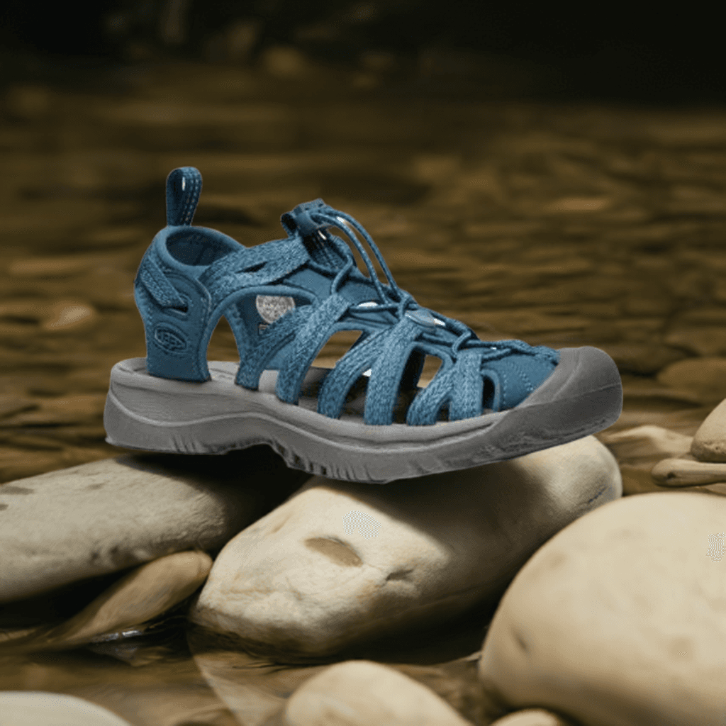 KEEN Whisper Fisherman Sandal in blue displayed on river rocks, emphasizing its outdoor-ready design and water-friendly materials.