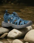 KEEN Whisper Fisherman Sandal in blue displayed on river rocks, emphasizing its outdoor-ready design and water-friendly materials.