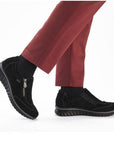Model wearing Women's Rieker 59071 Slip-On Shoe in black, styled with red pants, showcasing its casual waterproof wedge design.