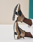 Brown and black wedge sneaker boots worn by a person sitting on a white surface with a beige and teal background.