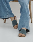 Woman in blue jeans sitting on wooden chair wearing blue knit slide sandals with tan outsole.