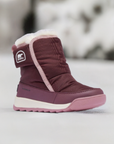 Maroon Sorel Kids Whitney II Plus Waterproof Snow Boots displayed in snowy outdoor setting, highlighting winter-ready design.