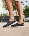 Navy blue boat shoes with brown accents worn by a person on a blurred outdoor background.