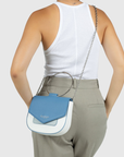 A woman shown from the back wearing a white tank top and light gray trousers, carrying a small crossbody bag with a blue flap, white body, and silver chain strap.