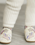 Child wearing white soft pants and leather soft shoes with unicorn and star designs.