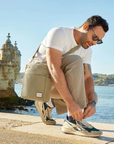 Man sitting by a waterfront with a beige bag, wearing sunglasses and a white shirt.