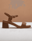 Side profile of brown Clarks Jaylan Ray Heeled Sandal with woven strap and block heel, styled against a warm-toned background.