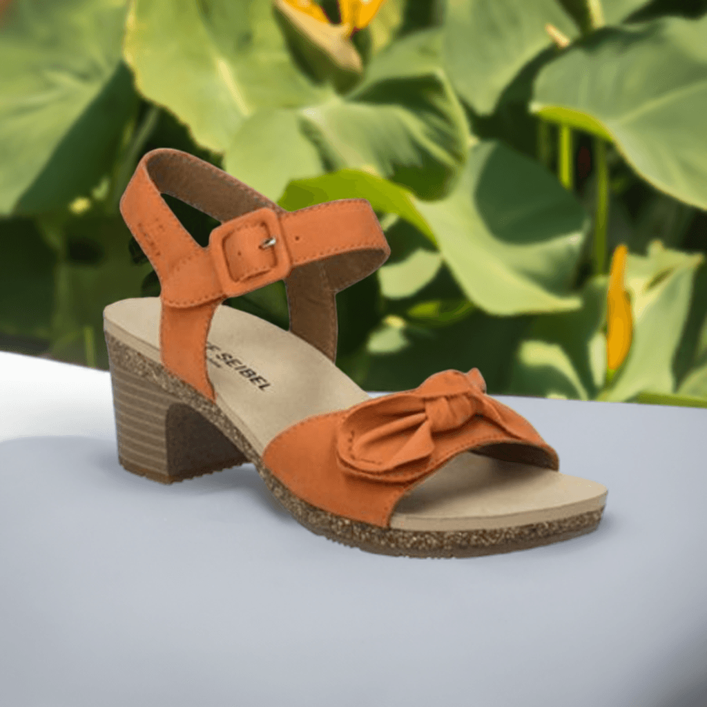 The Josef Seibel Grace 11 is a orange sling-back heeled sandal with a bow, featured on a lush green leaf background.