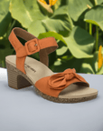 The Josef Seibel Grace 11 is a orange sling-back heeled sandal with a bow, featured on a lush green leaf background.