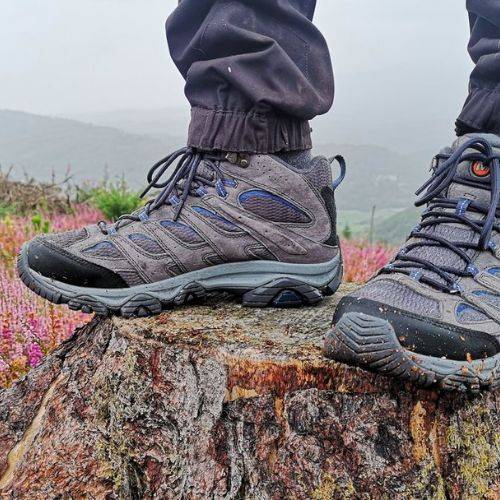 Women's Merrell Moab 3 Mid WP Boots in grey worn outdoors on a tree stump, showcasing waterproof hiking performance in rugged terrain.