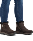 A pair of dark brown ankle winter boots with black faux-fur trim and side zippers, shown on a model wearing blue jeans.