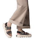 Camouflage-patterned platform sandals worn with beige pants.