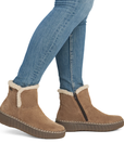 A person wearing tan suede ankle boots with white shearling lining and blue jeans, shown from the knees down.