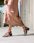 Person wearing a brown dress and black sparkly block heel sandals on a light-colored pavement.