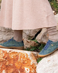 Woman in beige dress walking on rocks with green furry sneakers on.