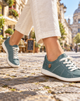 A person walking on a cobblestone street wearing teal blue leather sneakers with white laces and off-white cropped pants. The background shows a blurred outdoor urban street scene with buildings and a bicycle.