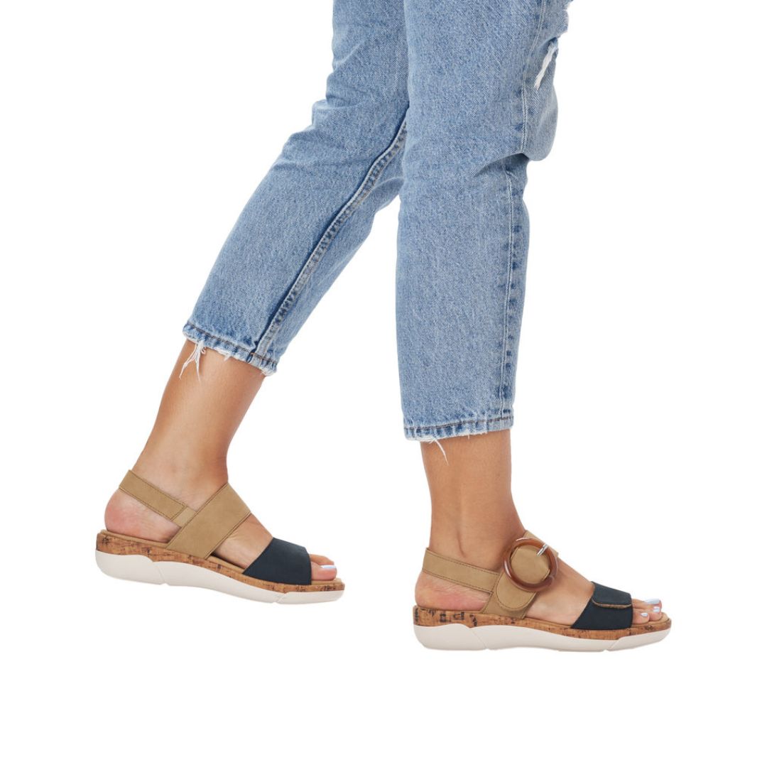 Model wearing Rieker R6853 Backstrap Sandal in tan and black, showcasing stylish oversized buckle and supportive flat sole for casual outings.