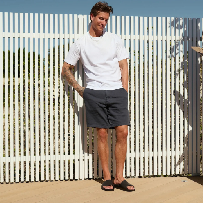 Man wearing black slide sandals, white t-shirt and dark shorts standing in front of a white picket fence.