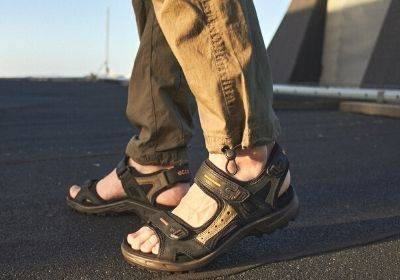 Person wearing brown outdoor sandals and rolled-up khaki pants standing on a rooftop with sunlight casting shadows.
