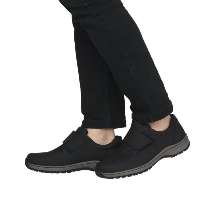 Legs in black pants wearing black shoe with Velcro closure and grey midsole.