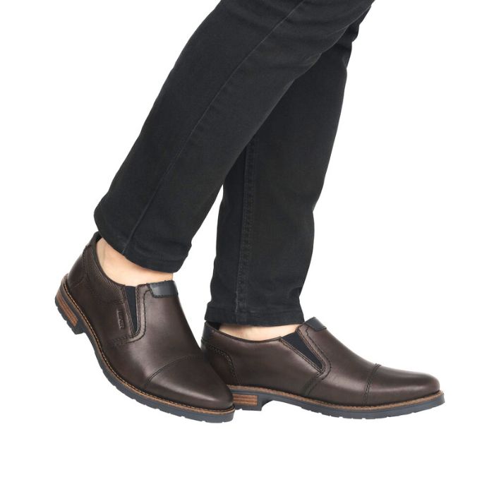 Men in black jeans wearing brown leather slip-on dress shoe with toe cap and navy accents and navy outsole.