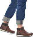 Legs in cuffed jeans wearing brown ankle boot with textile cuff, laces, white midsole and brown outsole.