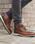 Legs in black jeans wearing brown ankle boot with textile cuff, laces, white midsole and brown outsole.