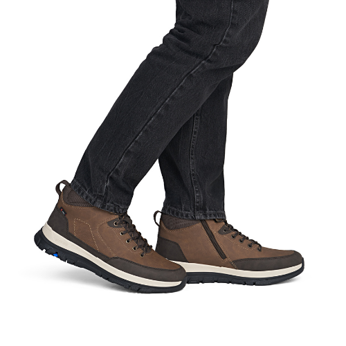 A person wearing brown mid-top lace-up shoes with dark jeans, shown from the calves down.