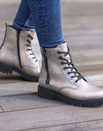 Legs in jeans wearing gold leather combat style boots with black laces, black lugged outsole and black outside zipper.