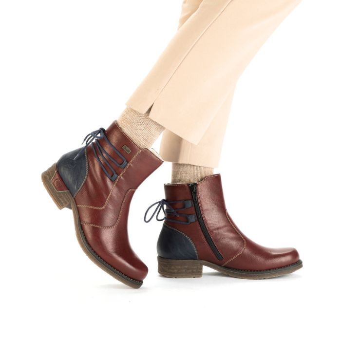Legs in tan pants wearing red leather ankle boot with low heel, navy heel counter and navy lace detail on heel.