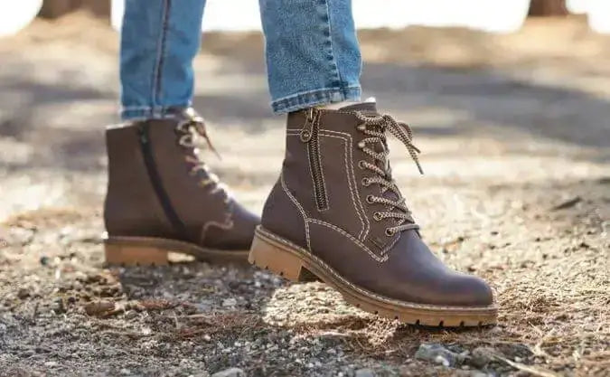 Brown leather lace-up ankle boots with rugged soles, worn with blue jeans, standing on a gravel and dirt outdoor path.