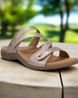 Gold strappy slide sandal sitting on a tan tree stump with trees blurred in background.