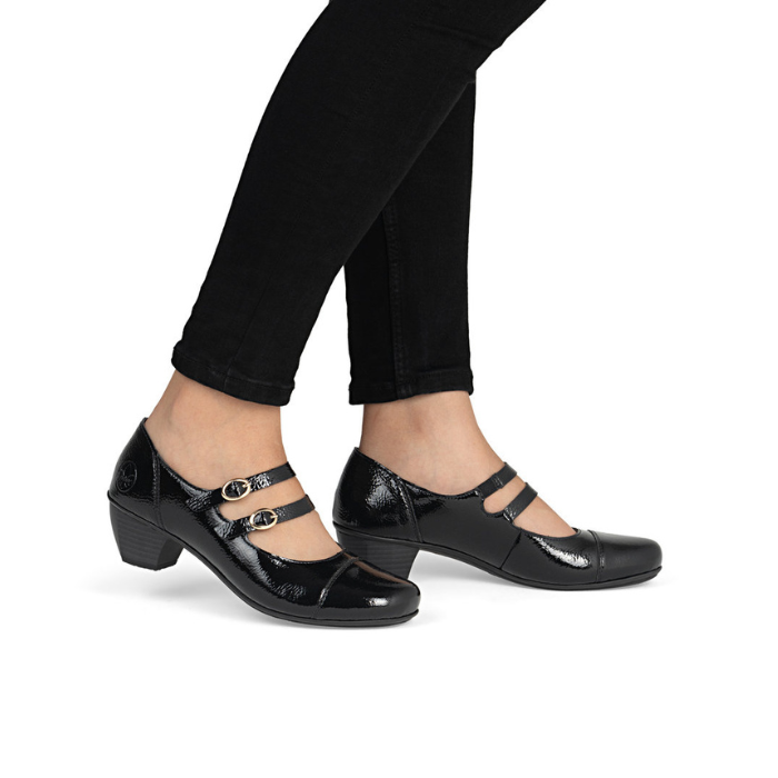 Black patent mary jane shoe with two gold buckle straps and a low block heel worn with black pants on model.